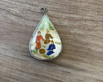 May include: Teardrop-shaped pendant with a miniature painting of three figures in colorful attire. The figures are set against a backdrop of a light green landscape. The pendant has a silver-colored metal frame and a small loop at the top for a chain.