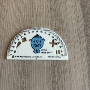 May include: A white plastic semi-circular protractor featuring a blue Little Miss Shy character illustration. The tool includes cutouts in the shapes of a diamond, a plus sign, and two vertical bars, with text reading Little Miss Shy, 1981 Roger Hargreaves, and 1985 Arby's, Inc.