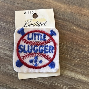 Puede incluir: Un parche cuadrado blanco con las palabras "LITTLE SLUGGER" en letras azules dentro de un diseño de béisbol rojo. El parche tiene acentos azules con temática de béisbol y está adherido a una etiqueta color crema con la palabra "Boutique" y el número "A-110".
