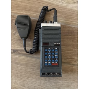 Puede incluir: Un transceptor de radio Yaesu portátil de color gris oscuro con un cable negro en espiral y un micrófono desmontable. El dispositivo cuenta con un teclado numérico con botones azules, una pequeña pantalla y varios interruptores etiquetados LOCK, UP y DISP.