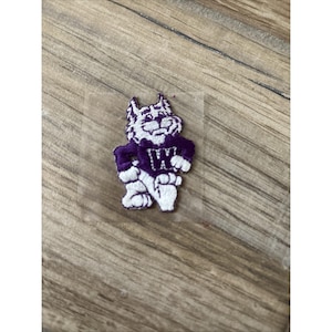 Op de afbeelding: Een geborduurde patch met een cartoon wildkat-mascotte in paarse en witte draad. De wildkat staat in een staande houding met de armen over elkaar en draagt een paars shirt met een opvallende witte letter W op het borstgedeelte.