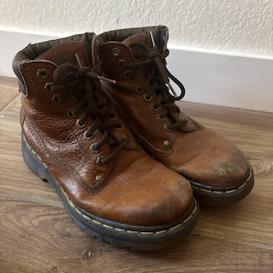Puede incluir: Un par de botas de cuero marrón desgastadas con cordones marrones oscuros y suela negra. Las botas tienen un aspecto robusto y desgastado, con desgaste visible en el cuero. Las suelas están cosidas con hilo amarillo.