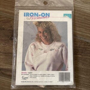 Puede incluir: Un kit de manualidades Iron-On Fashions de Golden Bee Stitchery que presenta una sudadera rosa claro con un diseño de conchas marinas. El kit incluye un patrón de transferencia termoadhesivo, hilo 100% algodón, pedrería, aguja e instrucciones.