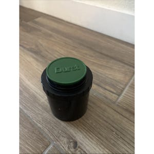 Może przedstawiać: Czarny zbiornik do wywoływania filmu z zieloną pokrywą. Na pokrywie wytłoczono słowo "Durst" w jasnym zielonym kolorze. Zbiornik jest cylindryczny i stoi na drewnianej powierzchni.