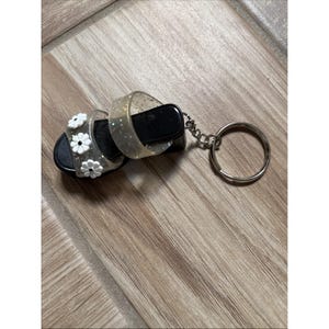 Puede incluir: Un llavero de sandalia en miniatura con suela negra, correas transparentes y detalles de margaritas blancas. Las correas transparentes están adornadas con purpurina. Un llavero de color plateado está unido a la sandalia.