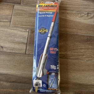May include: An Estes QCC Explorer flying model rocket kit in clear plastic packaging. The rocket is white with a red nose cone and black fins. The packaging displays text including 'Flies up to 1225 feet' and '35 inches tall' with a barcode at the base.