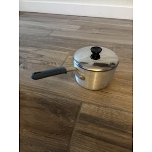 May include: A small, silver-colored saucepan with a lid and a black handle. The lid has a black knob. The handle is dark gray. The saucepan is sitting on a wooden surface.