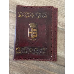 Peut inclure: Un porte-passeport en cuir bordeaux foncé orné d'un emblème en forme de bouclier doré avec une couronne et l'inscription STRESA. La couverture présente des motifs de volutes complexes et une bordure cousue avec des trous de laçage visibles.