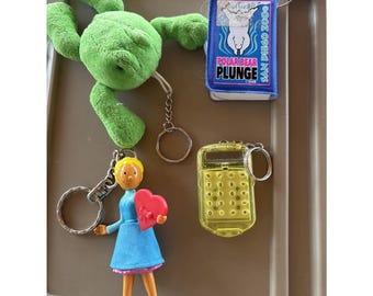 May include: A collection of four keychains: a green plush frog, a blue rectangular San Diego Zoo Polar Bear Plunge tag, a yellow translucent plastic calculator, and a figurine of a person in a blue dress holding a red heart-shaped box.