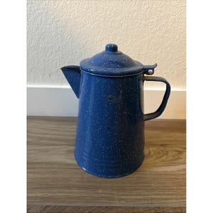 May include: A blue enamel coffee pot featuring a white speckled pattern throughout the exterior. The pot includes a matching lid with a small knob, a side handle, and a pointed spout. The finish is glossy with a visible metallic construction.