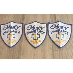May include: Three shield-shaped embroidered patches with white backgrounds and blue borders. Each patch features the text Mercy Ambulance in blue, a blue cross symbol with a yellow caduceus, and the text LAS VEGAS NEV. in yellow lettering.