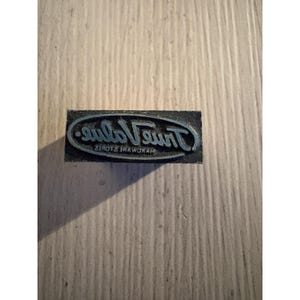 May include: A rectangular black metal printing block featuring the True Value Hardware Stores logo in raised relief. The design includes the text True Value inside an oval border with the words Hardware Stores printed underneath the main text.