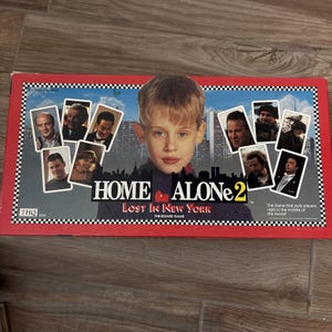 Puede incluir: Caja del juego de mesa Home Alone 2: Lost in New York. La imagen muestra al personaje principal rodeado de otros personajes. El título de la película se muestra en un fondo rojo.