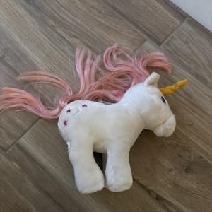 May include: A white plush unicorn toy featuring a long, flowing pink mane and tail. The unicorn has a yellow horn on its forehead and a small embroidered design on its flank consisting of a crescent moon surrounded by three small pink stars.