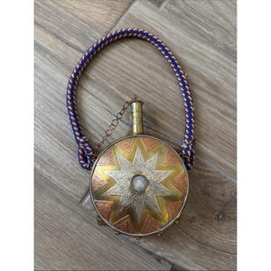 Puede incluir: Una petaca redonda de metal con un diseño de estrella central y paneles alternos en tonos oro y cobre. La petaca tiene un cuello cilíndrico pequeño con una cadena y un asa gruesa de cuerda trenzada en colores azul, rojo y blanco.