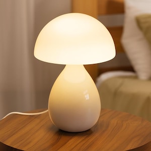 May include: A cream-colored mushroom-shaped table lamp with a rounded top and a teardrop-shaped base. The lamp is illuminated, casting a warm glow. The lamp is sitting on a wooden table.