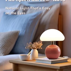 May include: A modern table lamp with a round, coral-colored base and a white, mushroom-shaped shade. The lamp sits on a wooden nightstand next to a small vase of flowers and a book. Text on the image reads: "Full-Spectrum LED | Ra98 Natural Light That's Easy on the Eyes."
