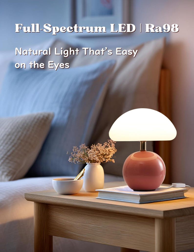 May include: A modern table lamp with a round, coral-colored base and a white, mushroom-shaped shade. The lamp sits on a wooden nightstand next to a small vase of flowers and a book. Text on the image reads: "Full-Spectrum LED | Ra98 Natural Light That's Easy on the Eyes."