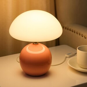 May include: A modern table lamp with a rounded, coral-colored base and a large, off-white, mushroom-shaped shade. The lamp is turned on, emitting a soft glow. The lamp is on a white surface.