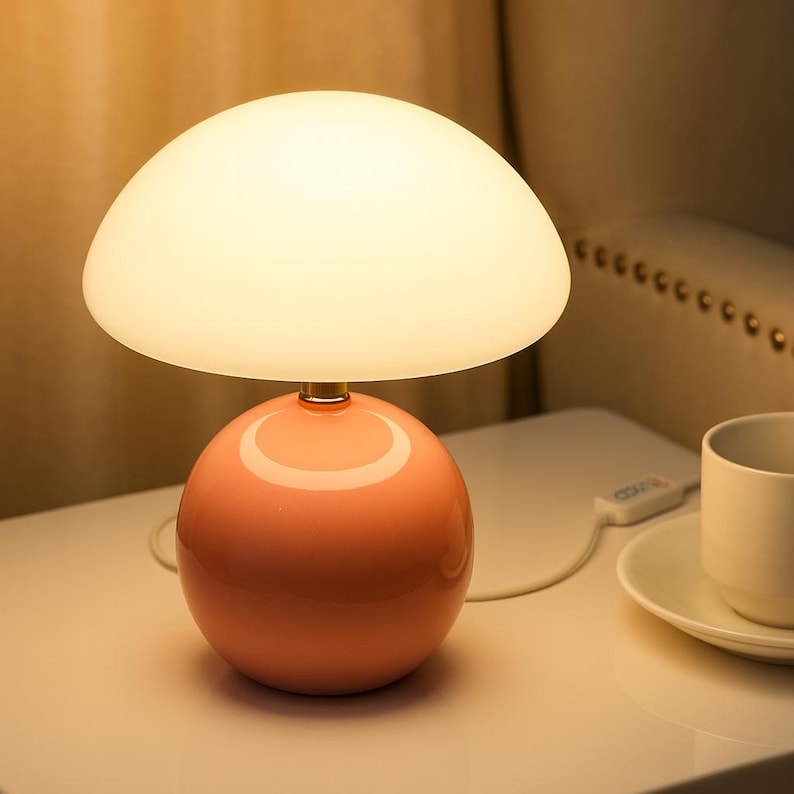 May include: A modern table lamp with a rounded, coral-colored base and a large, off-white, mushroom-shaped shade. The lamp is turned on, emitting a soft glow. The lamp is on a white surface.