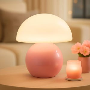 May include: A pink mushroom-shaped table lamp with a white, dome-shaped shade. The lamp sits on a light-colored wooden table, next to a lit candle in a glass holder and a small vase of pink flowers. The lamp's base is a sphere.