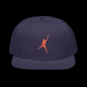 May include: A navy blue baseball cap featuring an orange silhouette of a person jumping and holding a ball. The cap has a flat brim and a structured crown, suitable for casual wear.