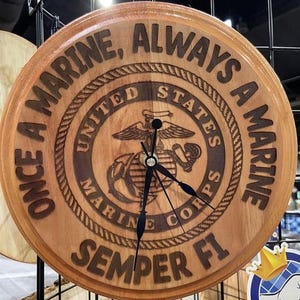 May include: A round wooden clock with the words "ONCE A MARINE, ALWAYS A MARINE" engraved around the edge. The center features the United States Marine Corps emblem and the words "SEMPER FI". The clock hands are black.