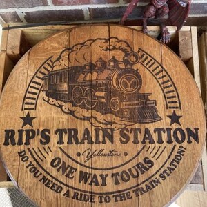 May include: A round, wooden sign with the text "RIP'S TRAIN STATION" and "ONE WAY TOURS" in a circular design. A detailed illustration of a steam train is in the center. The sign is made of light brown wood and has a rustic appearance.