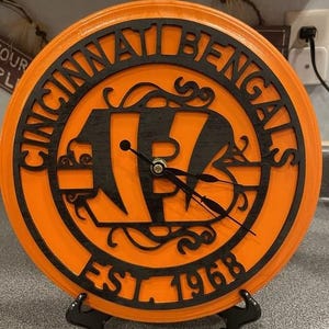 May include: Orange and black wooden clock featuring the Cincinnati Bengals logo and the text "CINCINNATI BENGALS EST. 1968". The clock has black hands and is displayed on a black stand. A decorative item for sports fans.