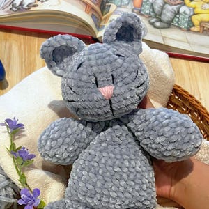Puede incluir: Un peluche gris con forma de rat&oacute;n, con una nariz rosa y detalles cosidos en negro. El juguete est&aacute; sobre una toalla blanca dentro de una cesta de mimbre. Un libro infantil est&aacute; abierto en el fondo.