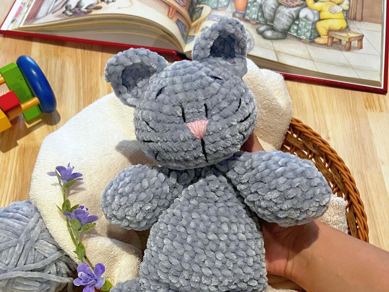 Puede incluir: Un peluche gris con forma de rat&oacute;n, con una nariz rosa y detalles cosidos en negro. El juguete est&aacute; sobre una toalla blanca dentro de una cesta de mimbre. Un libro infantil est&aacute; abierto en el fondo.