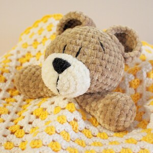 Puede incluir: Un oso de peluche hecho a mano, con hocico blanco y nariz negra, descansando sobre una manta de ganchillo amarilla y blanca. El oso es de color marr&oacute;n claro y tiene orejas peque&ntilde;as y redondas. La manta tiene un patr&oacute;n de granny square.