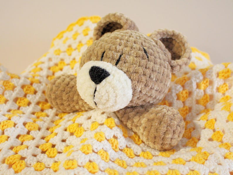 Puede incluir: Un oso de peluche hecho a mano, con hocico blanco y nariz negra, descansando sobre una manta de ganchillo amarilla y blanca. El oso es de color marr&oacute;n claro y tiene orejas peque&ntilde;as y redondas. La manta tiene un patr&oacute;n de granny square.