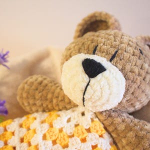 Puede incluir: Un oso de peluche tejido a mano con un cuerpo marr&oacute;n claro, hocico color crema y nariz negra. El oso tiene una manta estampada amarilla y blanca. La imagen tambi&eacute;n incluye flores moradas.