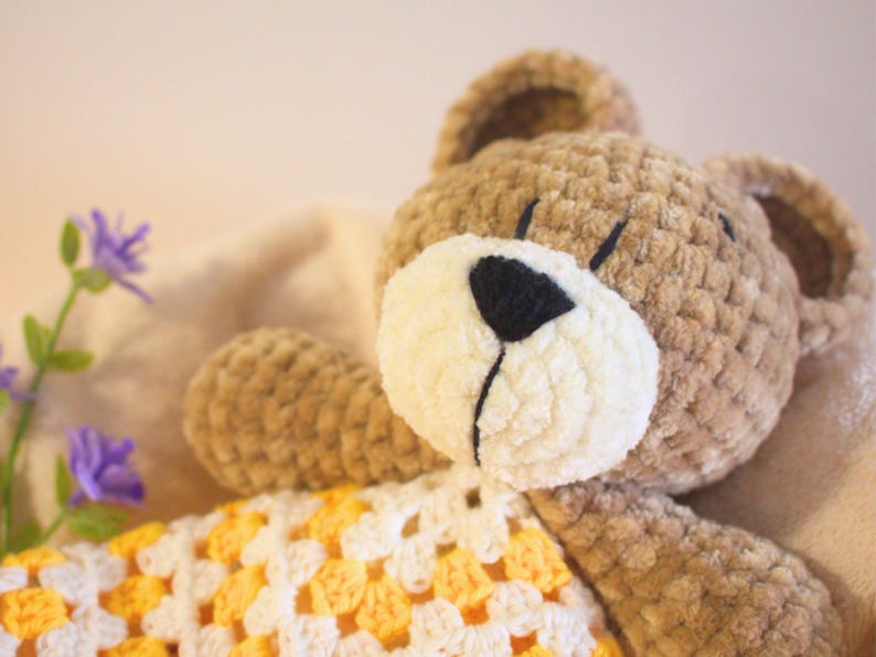 Puede incluir: Un oso de peluche tejido a mano con un cuerpo marr&oacute;n claro, hocico color crema y nariz negra. El oso tiene una manta estampada amarilla y blanca. La imagen tambi&eacute;n incluye flores moradas.