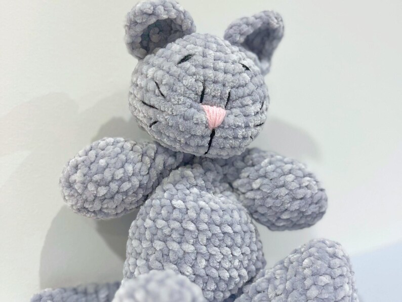 Puede incluir: Un peluche de gato gris hecho a mano con ganchillo, con una nariz rosa y detalles cosidos en negro. El gato tiene orejas grandes y est&aacute; hecho de hilo suave y texturizado. Un animal de peluche lindo y tierno.
