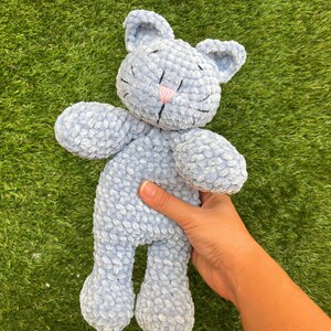 Puede incluir: Un peluche de gato azul claro con una nariz rosa y detalles cosidos en negro. El gato tiene orejas grandes y est&aacute; hecho de un material suave y texturizado. El juguete se sostiene sobre un fondo de hierba verde.