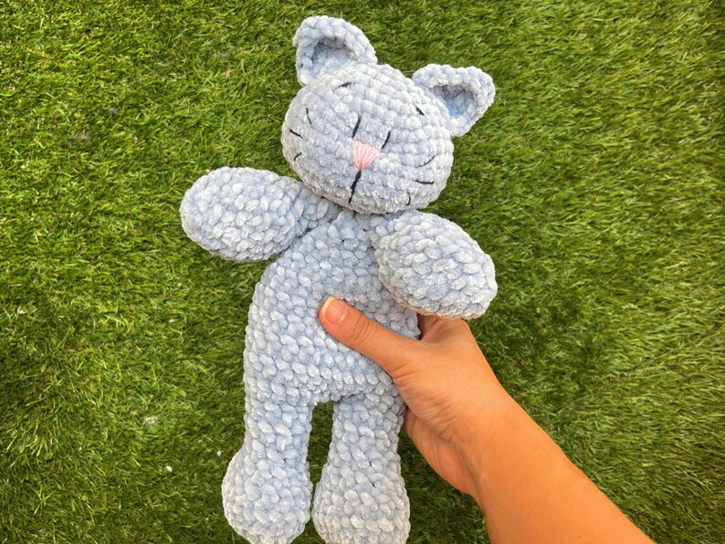 Puede incluir: Un peluche de gato azul claro con una nariz rosa y detalles cosidos en negro. El gato tiene orejas grandes y est&aacute; hecho de un material suave y texturizado. El juguete se sostiene sobre un fondo de hierba verde.