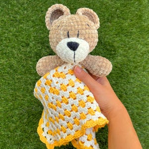 Puede incluir: Un oso de peluche hecho a mano, con cuerpo, orejas y extremidades de color marr&oacute;n claro. El oso tiene un hocico blanco y una nariz negra. Sostiene una manta de ganchillo amarilla y blanca. La manta tiene un patr&oacute;n de granny square.