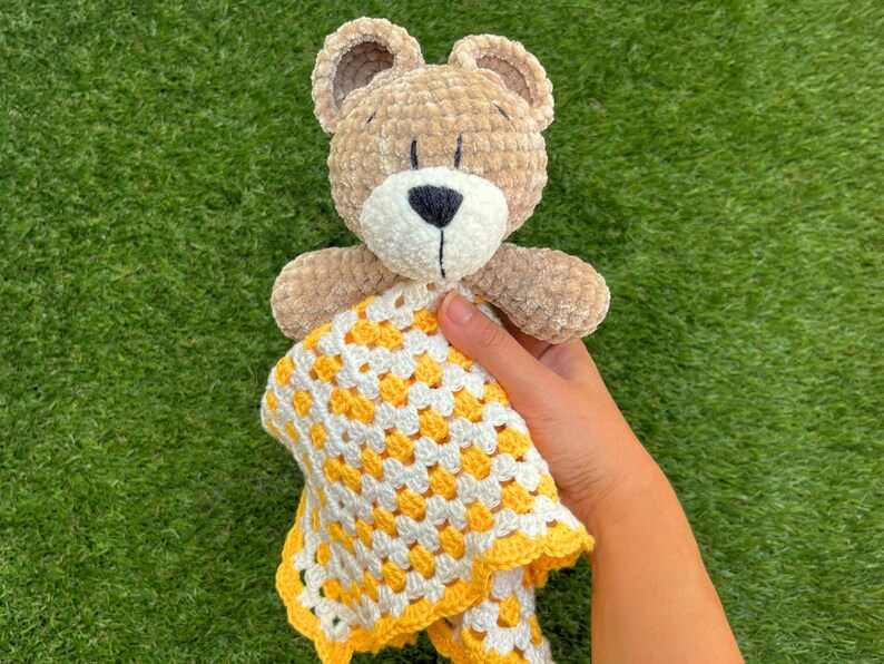 Puede incluir: Un oso de peluche hecho a mano, con cuerpo, orejas y extremidades de color marr&oacute;n claro. El oso tiene un hocico blanco y una nariz negra. Sostiene una manta de ganchillo amarilla y blanca. La manta tiene un patr&oacute;n de granny square.