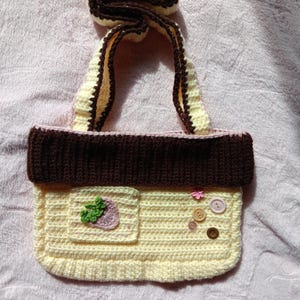 May include: A handmade crochet bag in cream, brown, and pink. The bag features a front pocket with a strawberry applique, decorative buttons, and a handle. The bag is a small tote style.