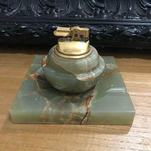 Peut inclure: Un briquet de table composé d'une base arrondie en onyx vert poli avec des veines brunes et d'un mécanisme d'allumage en métal doré, posé sur un plateau carré assorti en onyx vert doté de rainures intégrées pour le rangement.