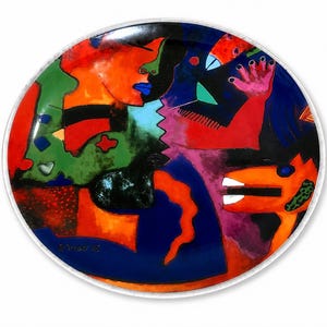 May include: A round ceramic plate featuring a vibrant, abstract composition with bold blocks of red, orange, green, blue, and black. The design includes stylized human-like profiles, a hand with fingers extended, and an animal figure with white teeth.