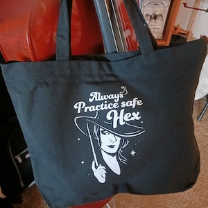 Puede incluir: Bolsa de tela negra con texto blanco que dice "Always Practice Safe Hex" y un gráfico de una bruja con un sombrero y una varita.