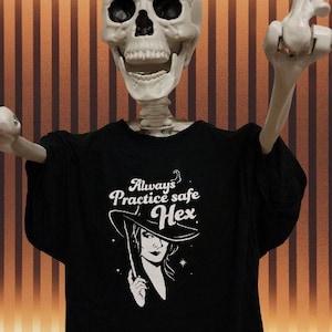 Puede incluir: Imagen temática de Halloween con un esqueleto de plástico que lleva una camiseta negra. La camiseta tiene el texto "Always Practice Safe Hex" con un gráfico de una bruja. El fondo tiene rayas verticales marrones y naranjas.