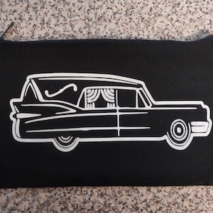 May include: A black canvas zippered pouch with a white outline of a vintage hearse. The hearse has a single window with a white curtain design.