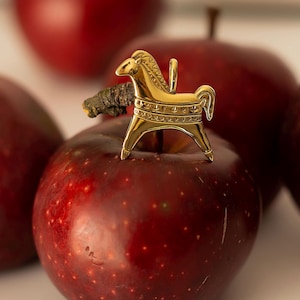 Op de afbeelding: Een goudkleurige paardenhanger met een lus, rustend op een rode appel. Het paard heeft gedetailleerde kenmerken, waaronder een manen en decoratieve patronen. De appel is dieprood van kleur, met een paar andere appels op de achtergrond.