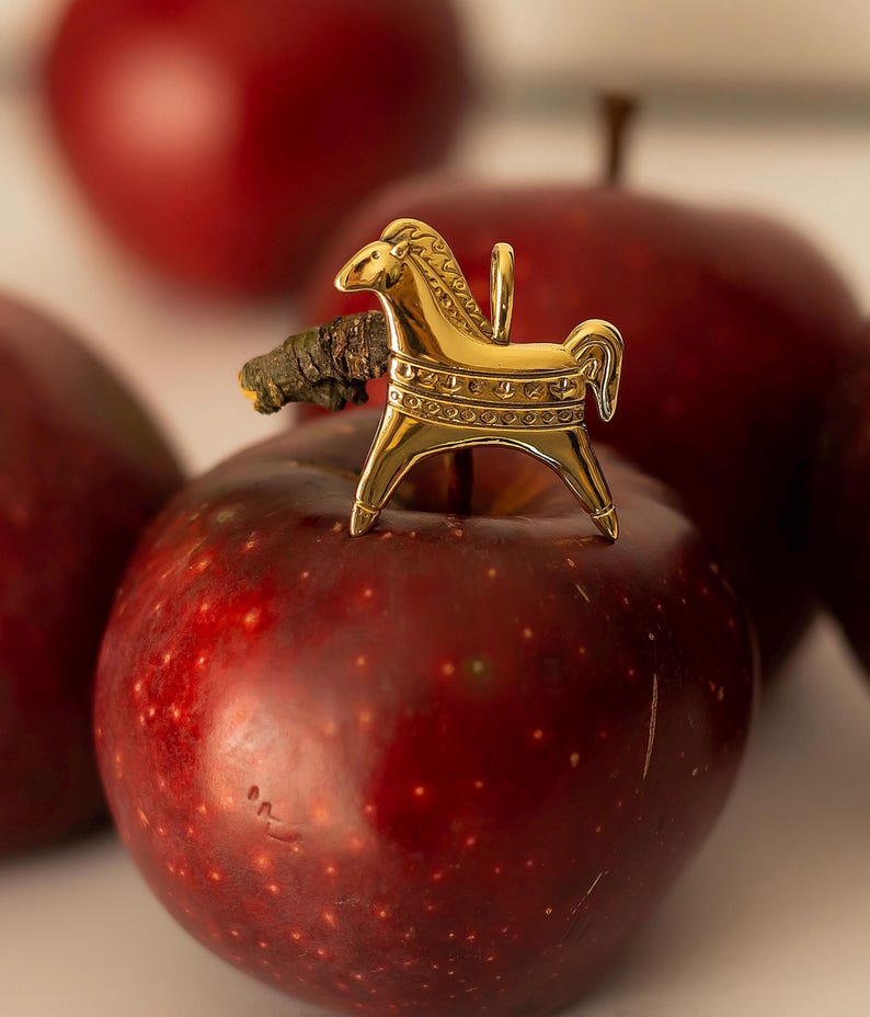 Op de afbeelding: Een goudkleurige paardenhanger met een lus, rustend op een rode appel. Het paard heeft gedetailleerde kenmerken, waaronder een manen en decoratieve patronen. De appel is dieprood van kleur, met een paar andere appels op de achtergrond.