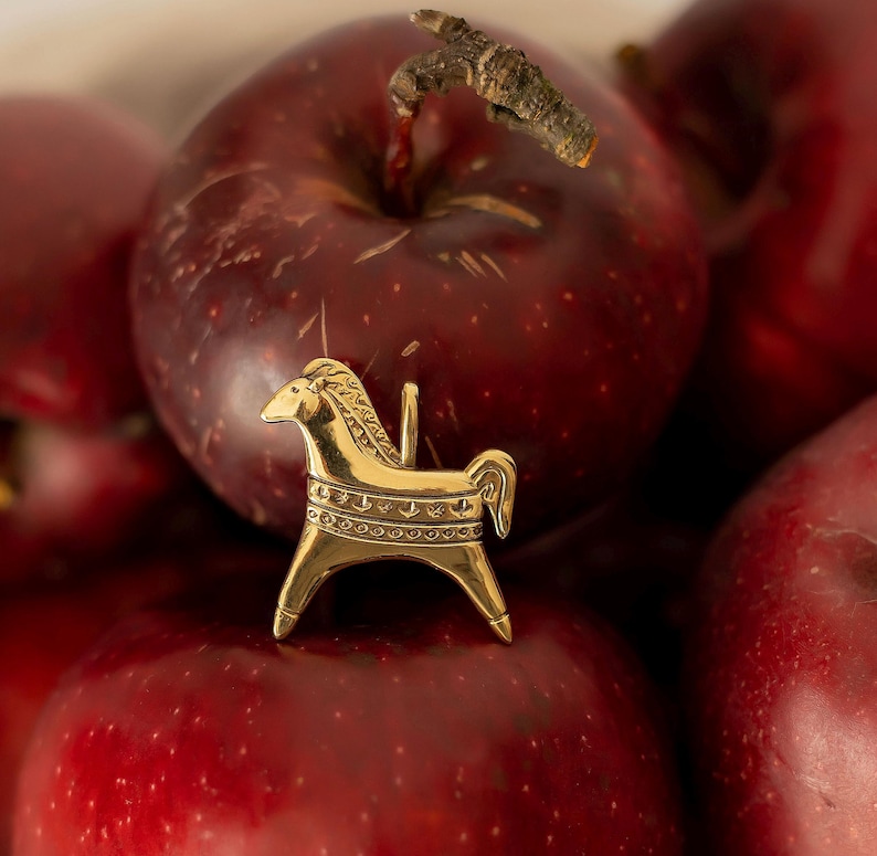 Op de afbeelding: Een kleine, goudkleurige paardenfiguur met ingewikkelde details, rustend op een bed van rode appels. Het paard heeft een gestileerde manen en staart, en is waarschijnlijk een decoratief item of bedel.