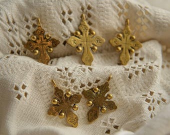 May include: A collection of antique gold-colored Orthodox crosses. The crosses feature intricate designs with small, round embellishments and religious inscriptions. They are displayed on a textured, off-white fabric.